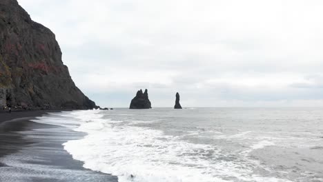 Foamy-ocean-waves-washing-upon-black-sand-beach-with-rock-sea-stacks