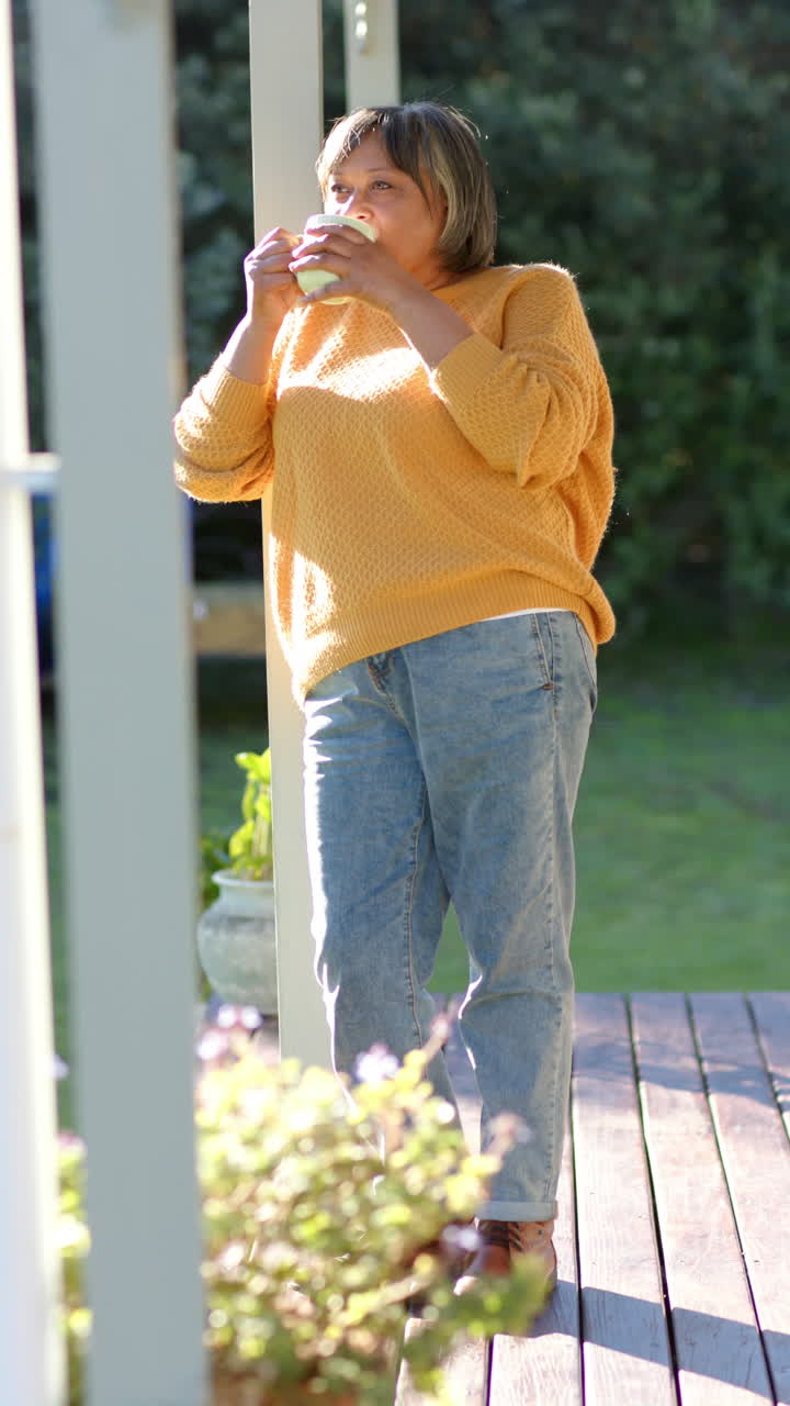 Vertical video of happy senior biracial woman drinking coffee on terrace, slow motion