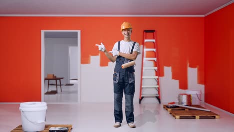 full body of asian man painter wearing safety helmet smiling and pointing to side while standing with painting wall in room