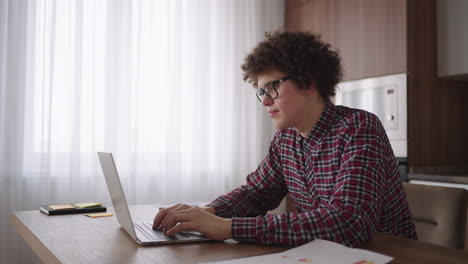 un hombre rizado con una mirada seria trabaja en una computadora portátil sentado en una cocina moderna. joven estudiante independiente usando computadora portátil estudiando en línea trabajando desde casa en internet