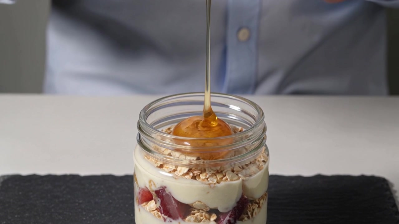 Tilting spoon person with watch in blue shirt pouring honey onto parfait on table