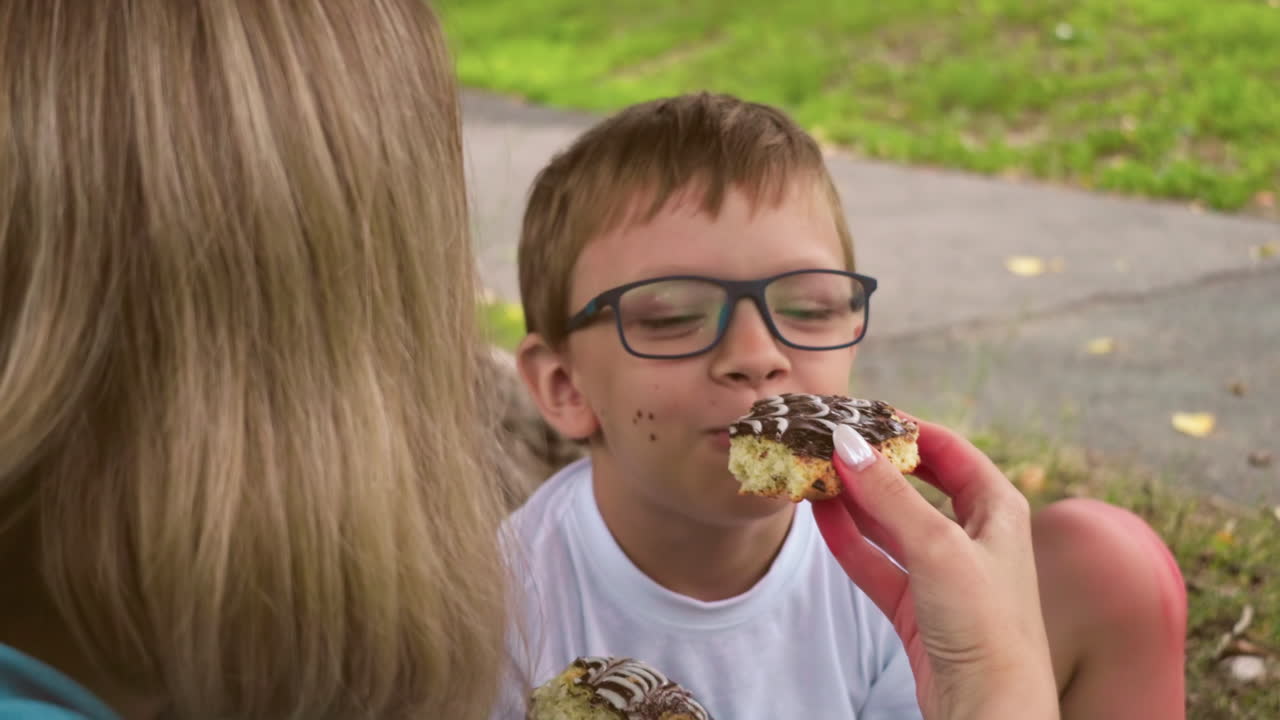 un momento lúdico entre una madre y su hijo, donde ella le ofrece un pastel de chocolate mientras está sentado en un campo cubierto de hierba bajo un árbol, el niño felizmente abre la boca para un bocado