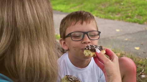 a playful moment between a mother and her child, where she offers him a chocolate cake while seated on a grassy field under a tree, the boy happily opens his mouth for a bite