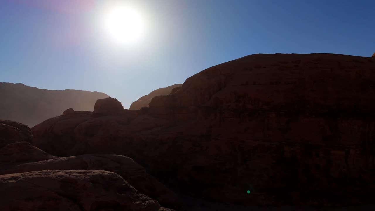 Uma sequência deslumbrante de drone em ângulo alto, capturando os acidentados picos de arenito de Wadi Rum, silhuetados pela intensa luz do dia e reflexo da lente.