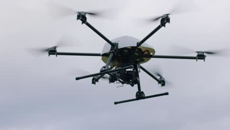 Handheld-camera-shot-of-black-and-yellow-drone-with-six-propellers-and-a-camera-in-flight-against-a-cloudy-sky