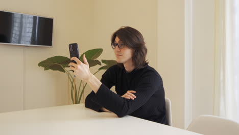 Young-man-with-glasses-on-a-video-call-using-his-mobile-phone,-sitting-at-a-table