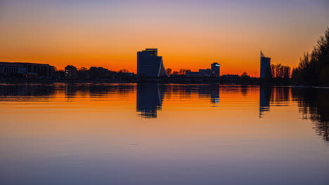 time lapse shot of tranquil lake during golden sunset and silhouette of national library of latvia in background - riga,europe