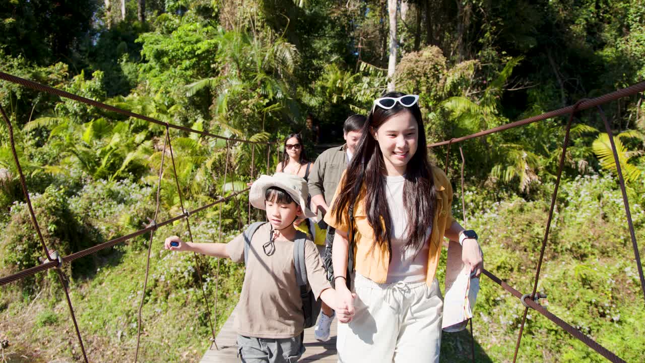 Família feliz atravessa ponte suspensa na floresta durante uma caminhada ensolarada de férias, câmera portátil rastreando para frente