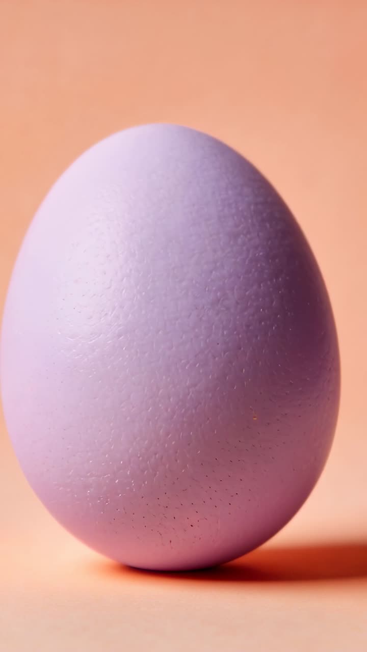 Vertical video: Tilting lilac egg leaning and rolling down peach table after tilt, showing shadow