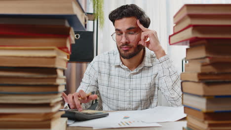 exhausted indian businessman reviewing documents reports using calculator working hard in office