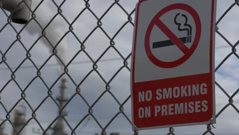 a refinery pipe smoking and polluting in the background pulling focus to a no smoking sign in the foreground on a chain-linked fence