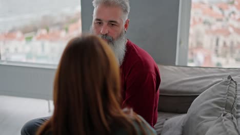 Over-her-shoulder,-a-brunette-girl-in-a-green-jacket-measures-the-temperature-with-a-thermometer-of-her-elderly-dad-with-gray-hair-and-a-lush-beard-in-a-red-T-shirt-Sitting-on-the-sofa-in-a-modern-apartment-overlooking-the-sea