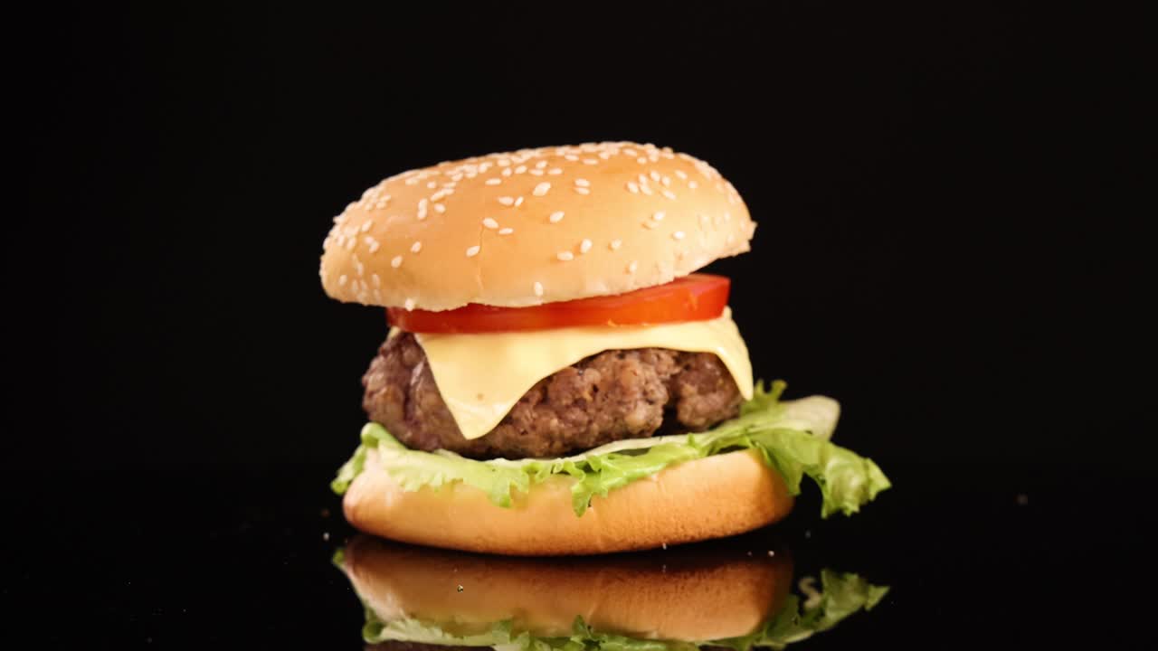 Golden French fries fall onto a cheeseburger with lettuce, tomato, and sesame bun, arranged on a reflective surface with dramatic studio lighting and black background
