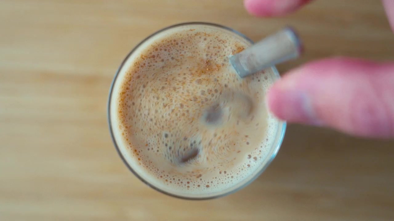 Primo piano diurno di una mano che mescola un chai latte freddo con latte d'avena in un bicchiere con una cannuccia su una superficie di legno.