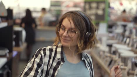 portrait of cute caucasian pretty young woman with short curly hair, plaid shirt and in headphones dancing and listening music in the middle of the supermarket with household stuff. slow motion