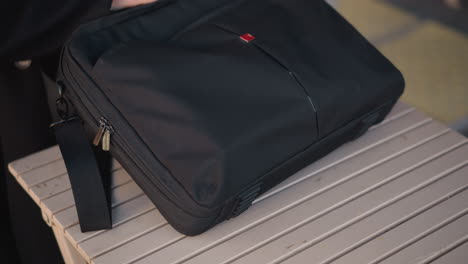 close-up of someone in black coat dropping a bag on a wooden table and unzipping it, showcasing white polished nails in a blurred urban outdoor setting