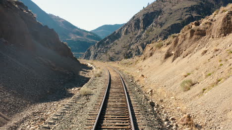 Aerial-down-desert-train-tracks-revealing-dramatic-valley-river-gorge-below