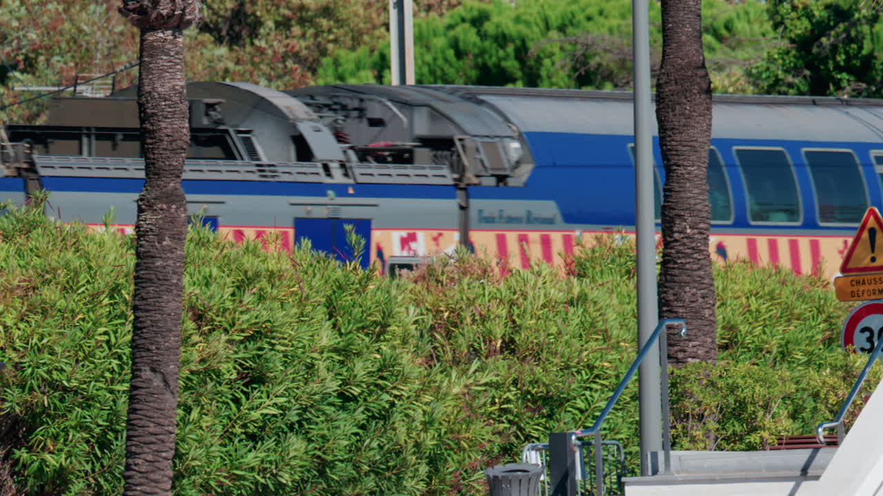 Cannes, France - October 7, 2025: A blue regional train passes near palm trees and a street