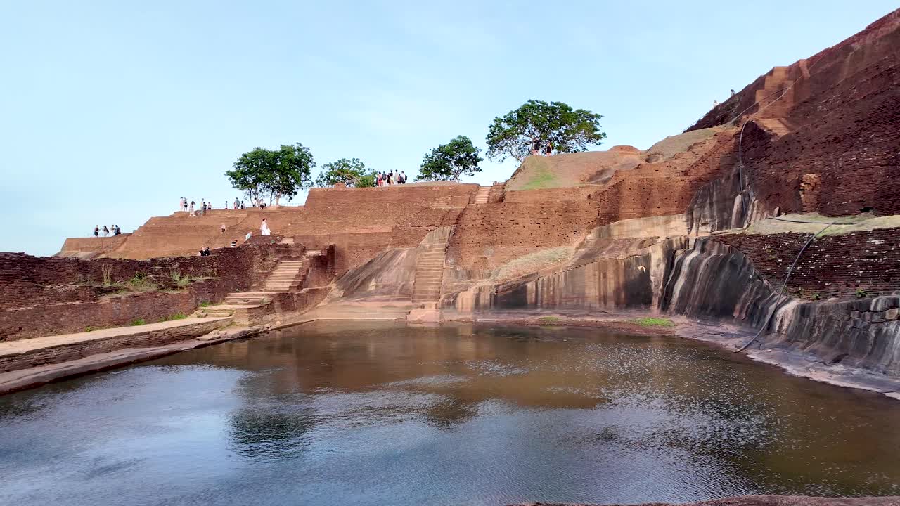 Ancient stone structures against a clear sky, with a tranquil water feature in the foreground, showcasing historical architecture and natural beauty.