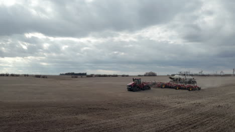 Large-red-industrial-seeding-machine-followed-by-fertilizer-in-dusty-farm-in-rural-countryside,-above-aerial-profile