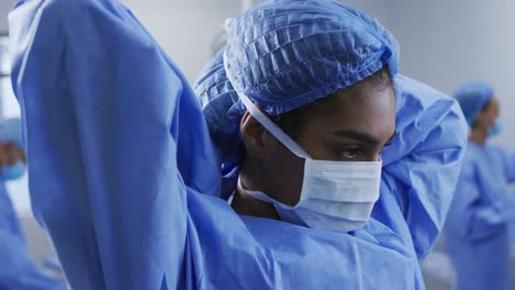 Mixed-race-female-surgeon-putting-on-protective-clothes-standing-in-operating-theatre