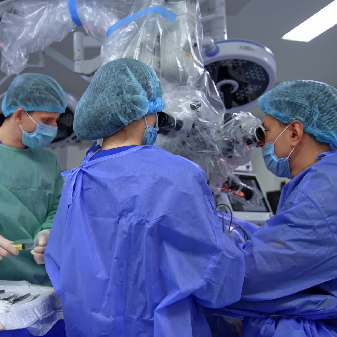 Male neurosurgeon and his assisting nurse looking at microscope at operation. Rear view of busy medical specialists carrying out the surgery