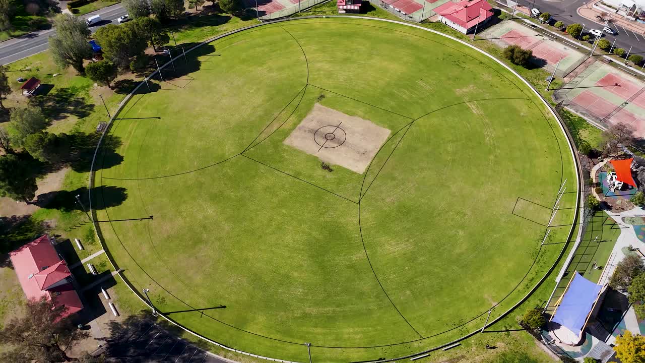 Drone camera circles above a large, empty sports ground with soccer goals and cricket pitch, under clear skies and bright natural daylight