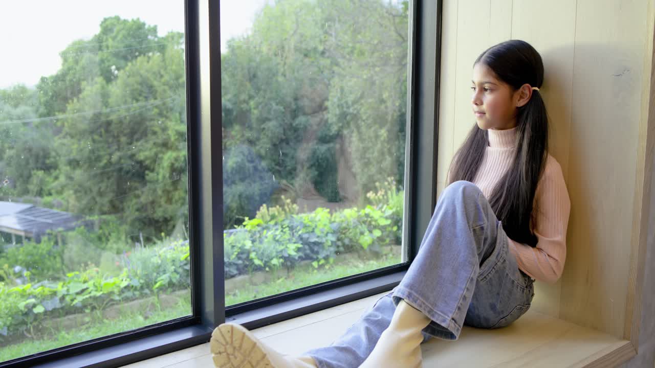 Panoramica e zoom della camera su una ragazza indiana seduta su una panca vicino alla finestra che guarda fuori, spazio per il testo