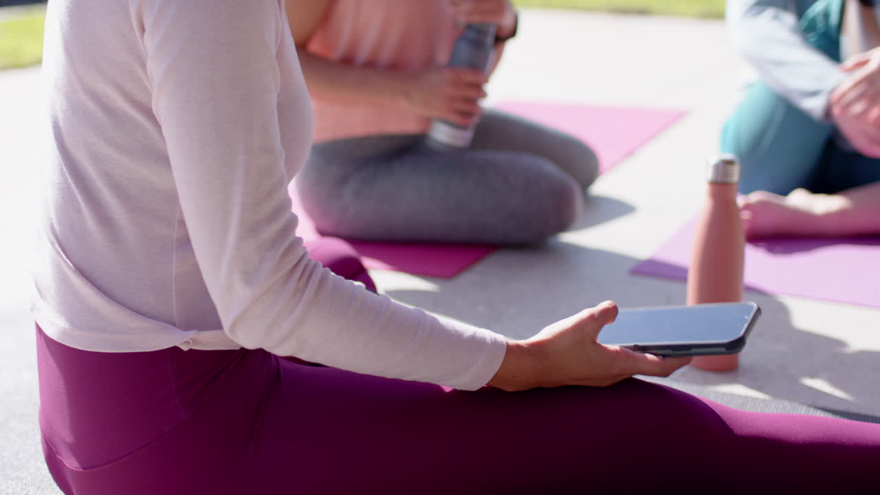 Using smartphone, woman sitting with female friends on yoga mats, enjoying outdoor hangout