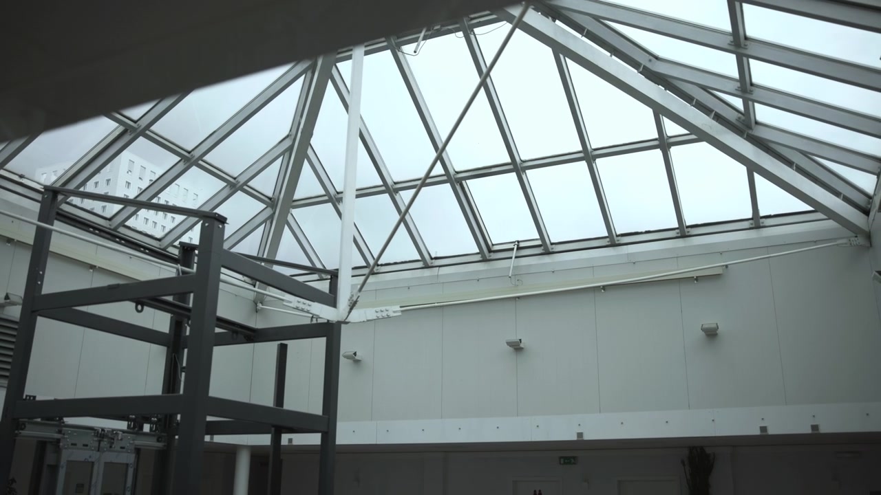 Upward interior view of modern atrium featuring glass skylight roof, steel support beams, pale wall panels, dark metal framework, and soft daylight across spacious geometric architecture