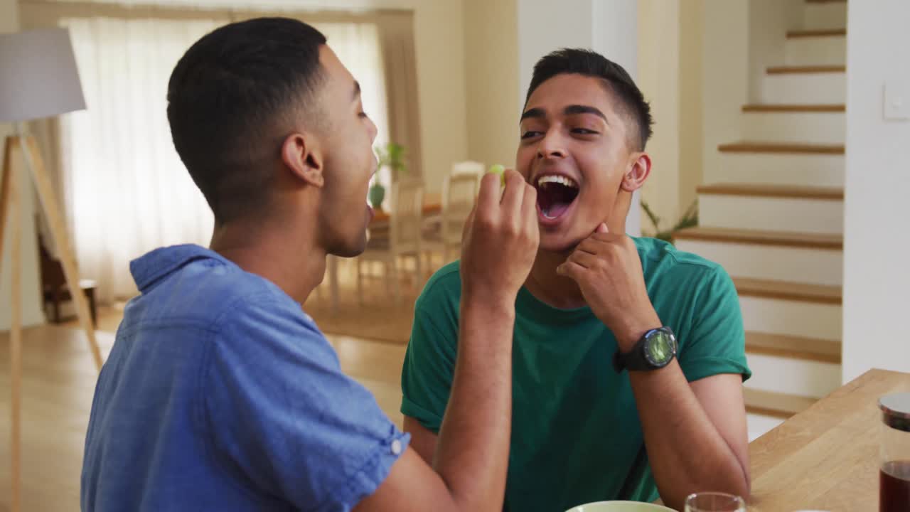 Smiling mixed race gay male couple having breakfast together in kitchen one feeding the other grapes