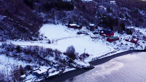 Vista-Aérea-De-Cabañas-Nevadas-En-La-Ladera-En-Invierno-Con-Mar-En-Calma-En-Noruega