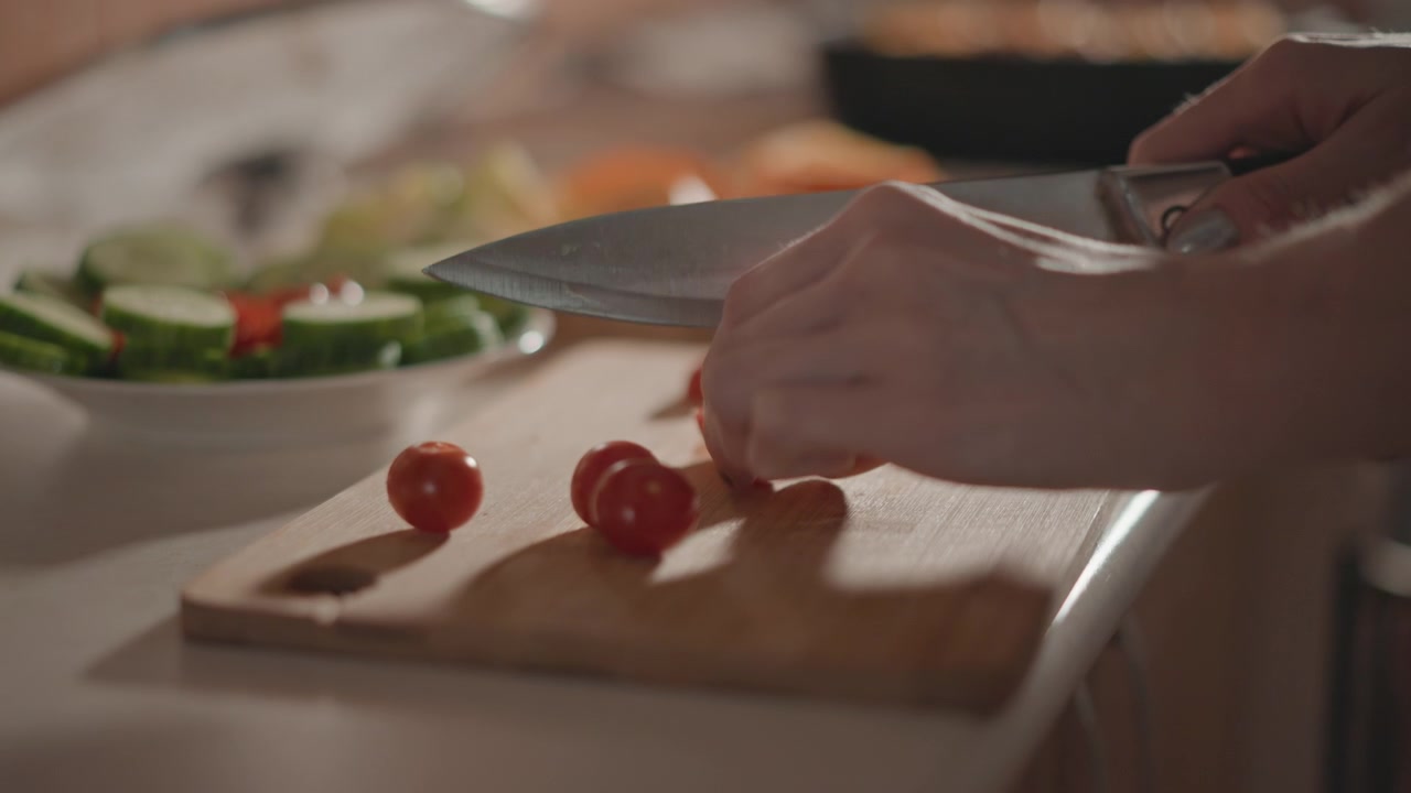 Primo piano di mani che affettano pomodorini su un tagliere di legno in una luminosa cucina domestica, con una ciotola per insalata e ingredienti freschi leggermente sfocati sullo sfondo, mostrando un'attenta preparazione del cibo e prodotti freschi.