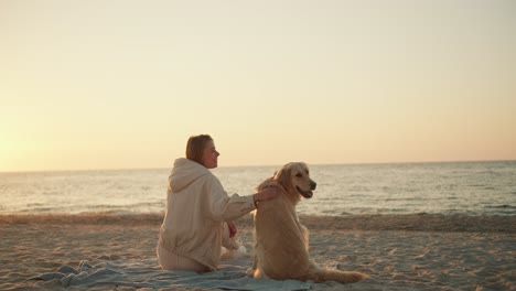 rear view of a blonde girl who sits next to her dog and interacts with her on the background of the beach and the sea in the morning