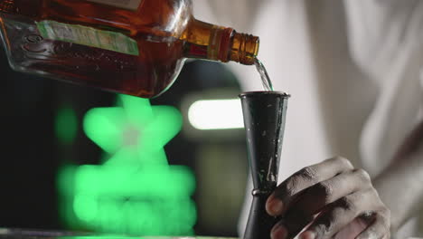 Bartender pouring whiskey from bottle into jigger for cocktail preparation, with focused expression and green neon lighting in background, creating a vibrant nightlife scene