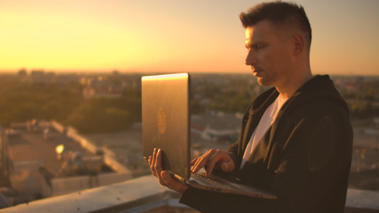 un programador con una computadora portátil se sienta en el techo y escribe código al atardecer. trabajo remoto freelance. libertad para trabajar. escribir en un teclado al atarrear el sol con una vista de la ciudad.