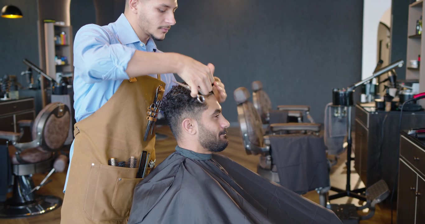 To voksne afrikanske mænd i en salon, hvor frisøren tager saks og kam for at klippe kundens hår og forme det.