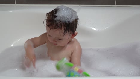 cute ukrainian-korean baby girl toddler playing with colorful toy turtle in the bath tub with foam