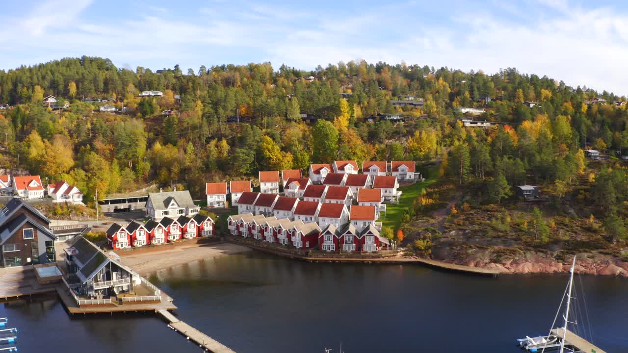 A drone reveals the little port of Holtnesstøa village, surrounded by calm waters and colorful houses. Some trees remain tinged with orange, capturing the charm of an autumn day.