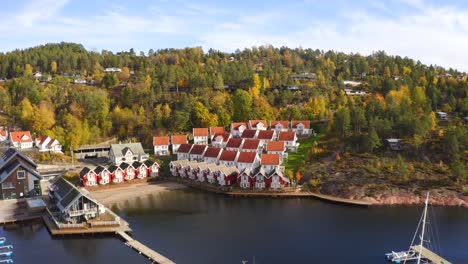 A drone reveals the little port of Holtnesstøa village, surrounded by calm waters and colorful houses. Some trees remain tinged with orange, capturing the charm of an autumn day.