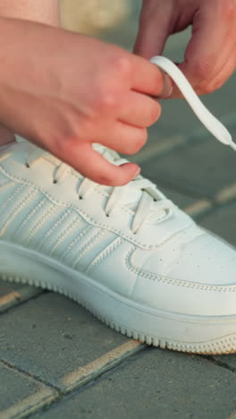 primer plano de una persona atando los cordones de zapatillas blancas en un camino de pavimento entrelazado, las manos ajustando cuidadosamente los cordones para un ajuste seguro, la toma detallada hace hincapié en el movimiento de la mano