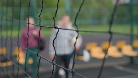 close-up of volleyball net in sharp focus with blurred view of athletes entering court in background, surrounded by greenery, capturing depth and anticipation for training or competition