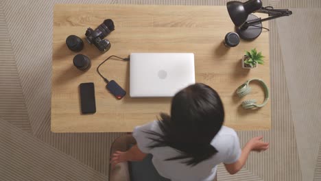 top view of asian woman editor walking into the workspace sitting down and using a laptop next to the camera editing the video at home