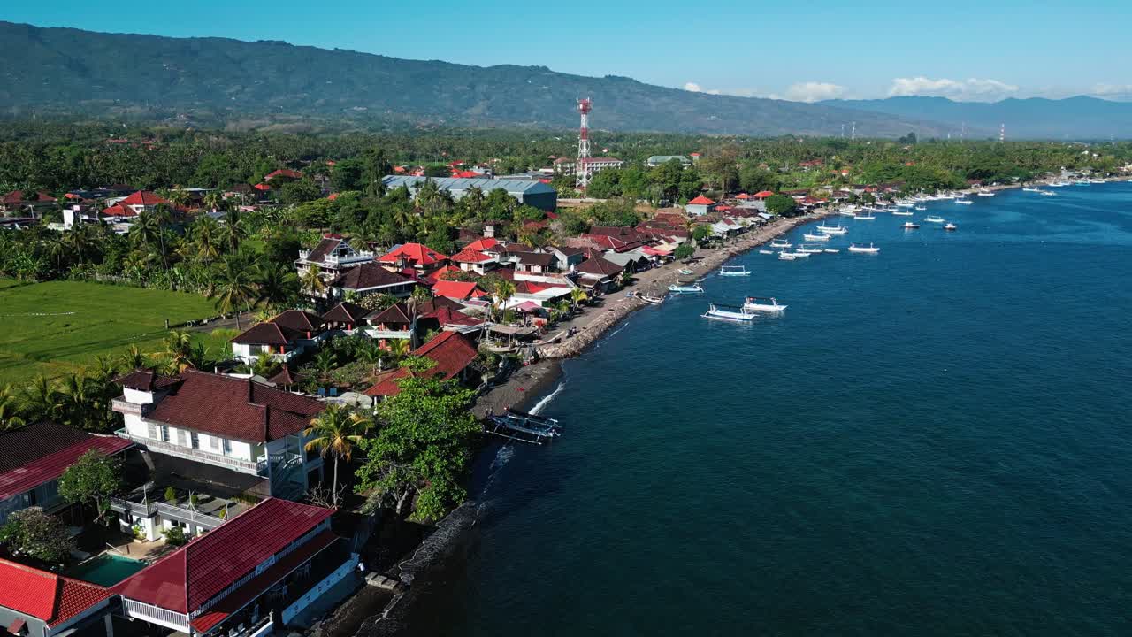 Drone aerial footage of North Bali coastline showing gentle waves, traditional fishing boats, houses, and lush tropical trees under bright daylight in a popular coastal tourist area