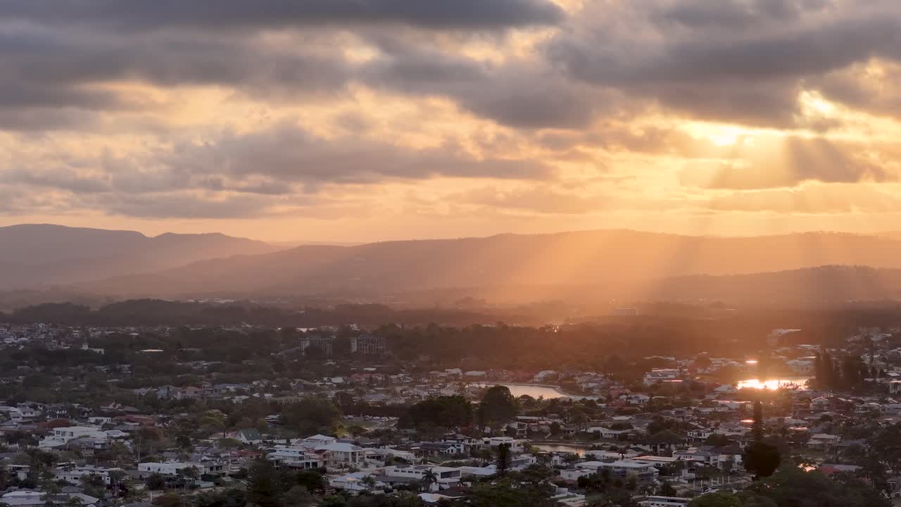 Dramatic sunset light beams through clouds over waterfront suburbs, wide aerial view, tranquil mood