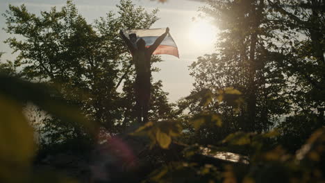 Perspectiva-De-Ojo-De-Rana-Sobre-El-Hombre-Que-Sostiene-La-Bandera-Eslovena-Hacia-El-Sol-De-La-Mañana-En-La-Cima-De-La-Montaña-Slivnica,-Entre-Los-árboles