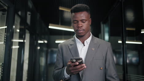 A-black-man-in-a-business-suit-walks-down-the-corridor-of-the-office-looking-at-his-mobile-phone.-Business-men-goes-with-a-phone-in-hand