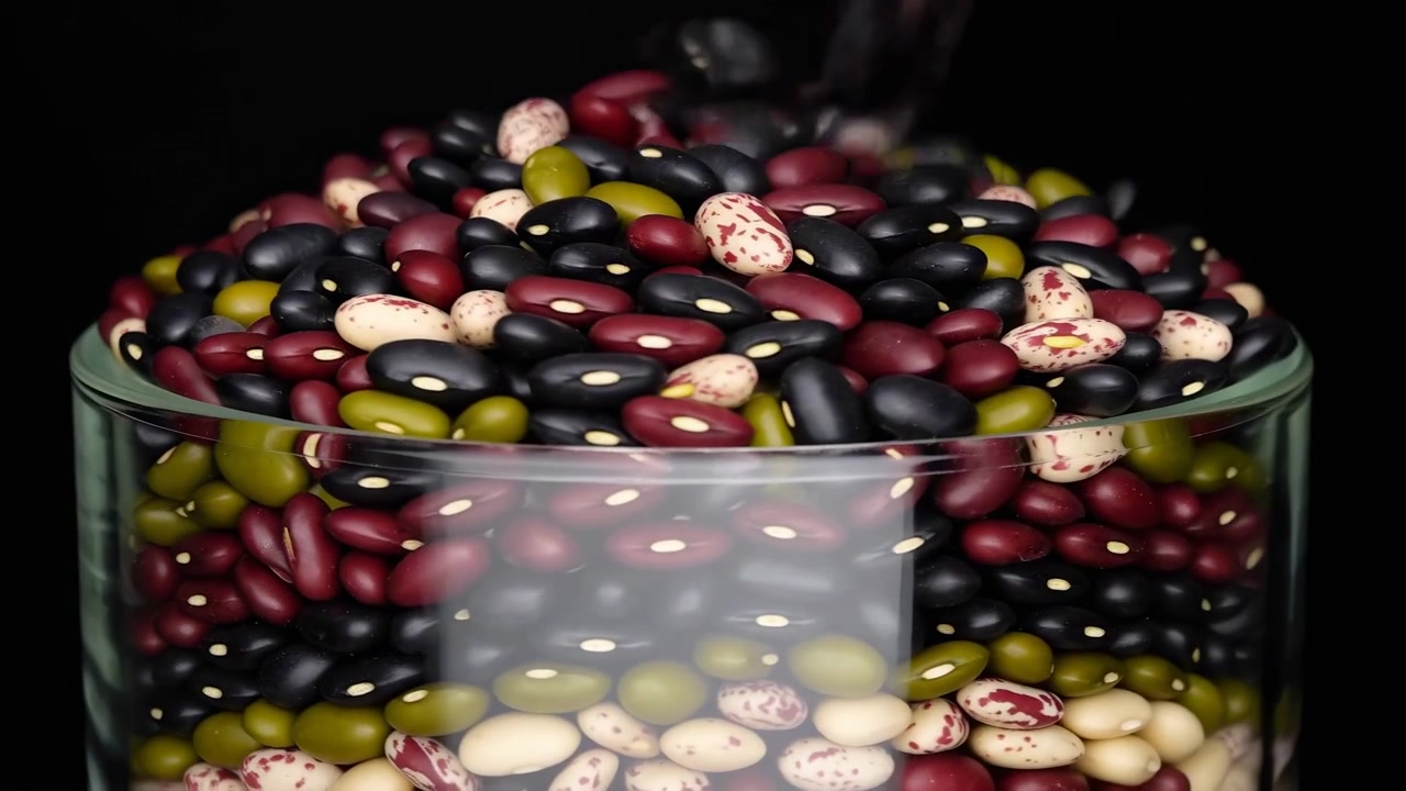 Mixed beans continue filling transparent glass container as individual legumes accumulate near top, showing varied shapes, colors, textures, and glossy surfaces under lighting in close composition.