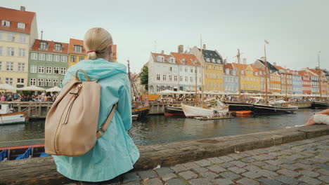 a woman sits on the embankment and admires the colorful buildings on the banks of the nyhavn canal i