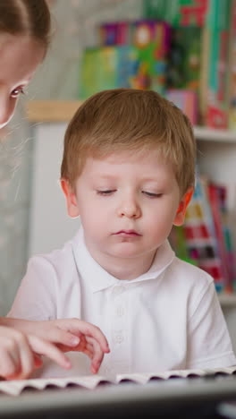 little girl makes smile on brother face with fingers trying to change mood during music lesson. sister motivates toddler boy to learn playing piano